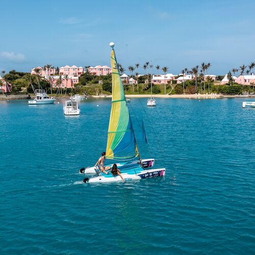 A small sailboat with colorful sails glides on a blue body of water, surrounded by other boats and a scenic shoreline with pink buildings.
