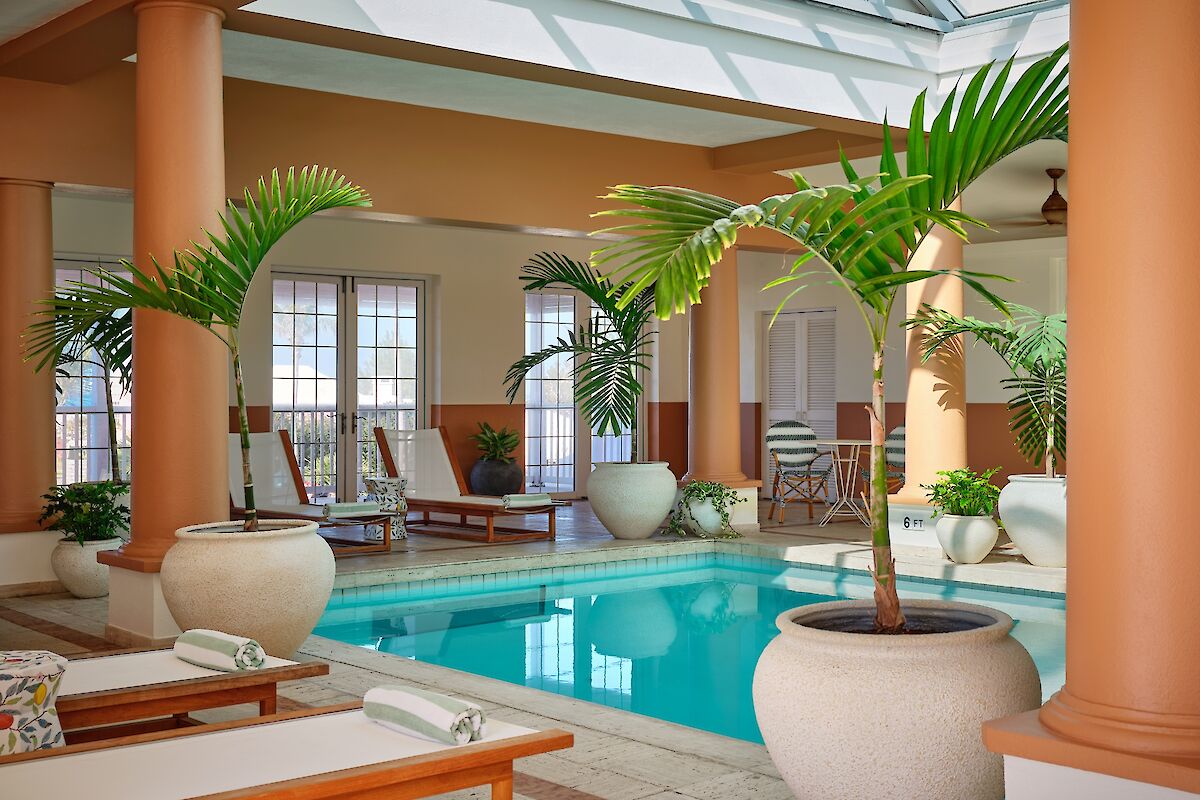An indoor pool area with orange columns, lounging chairs, tropical plants, and potted palms around a bright, tranquil pool.