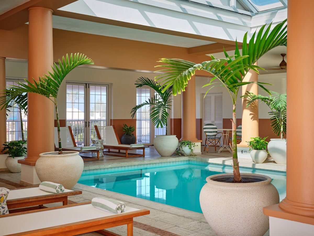 An indoor pool area with orange columns, lounging chairs, tropical plants, and potted palms around a bright, tranquil pool.