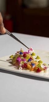A hand is plating a gourmet dish with colorful garnishes on a white rectangular plate.