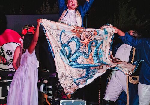 A theatrical performance with a person holding a fabric featuring an octopus design; others in costumes are gathered on stage.