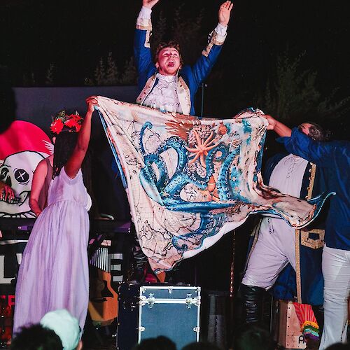 A theatrical performance with a person holding a fabric featuring an octopus design; others in costumes are gathered on stage.