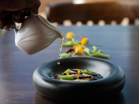 A chef pours a golden sauce from a glove into a bowl topped with vegetables and flowers.