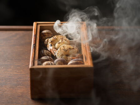 A wooden tray of roasted chicken and shellfish, steaming hot with fragrant smoke rising from the food.