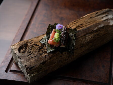 A plated sushi-like bite of pink fish and dark seaweed on a wooden board, topped with herbs and a purple edible flower.