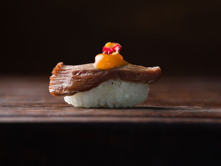 A sushi nigiri sits on a dark wooden surface, topped with tuna, a quail egg, and a small red garnish.