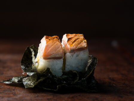 Two pieces of salmon nigiri sushi on a seaweed strip, resting on a wooden board in moody lighting.