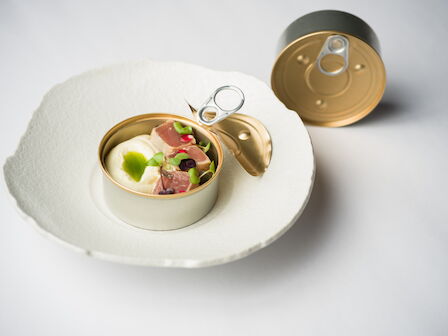 An open can of gourmet food on a plate, with the pull-tab opener, garnished with meat and fresh greens.
