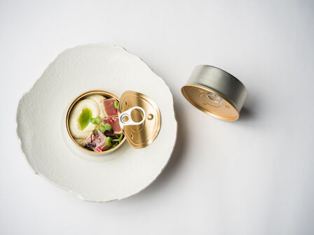 An opened gold can of pet food sits on a white plate, with visible chunks and bits of greens.