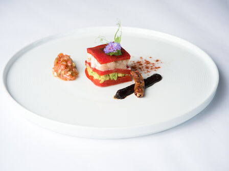 A plated gourmet dish: a red-capped seafood stack with green garnish, purple flower, dark vanilla bean, and a side bite.