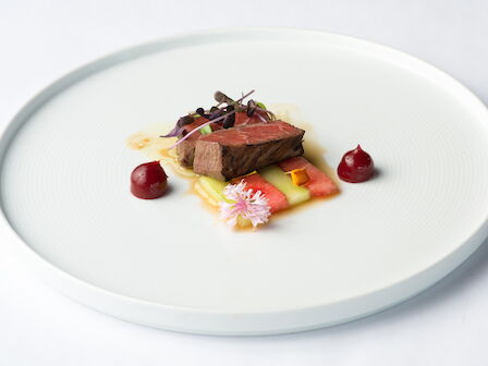 A plated steak with watermelon, herb sprouts, microgreens, and a bright sauce on a white dish.