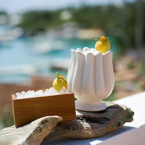 A beachside cocktail display: lemon-shaped garnishes on ice in metal cups with the ocean blurred behind them.