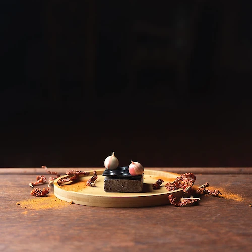 A dessert with chocolate, decorative elements, and two small spheres is on a round wooden plate on a wooden surface.