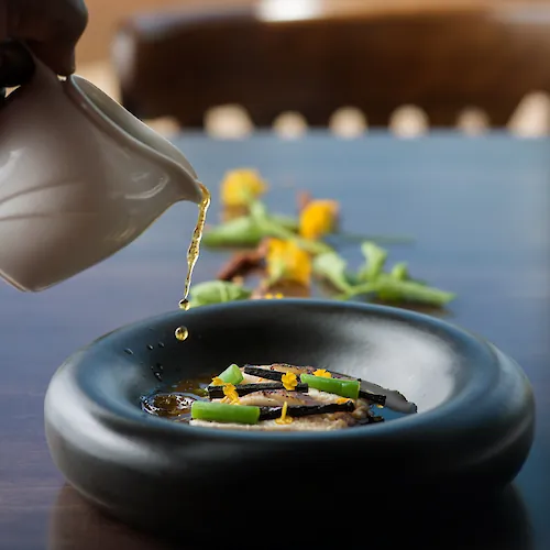 A gloved hand pours a golden oil sauce into a bowl of food with colorful vegetables in the background.