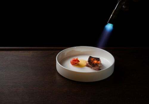 A lit torch sears a plated piece of steak with sauce and tomato on a white bowl against a dark background.