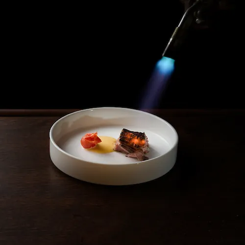 A lit torch sears a plated piece of steak with sauce and tomato on a white bowl against a dark background.