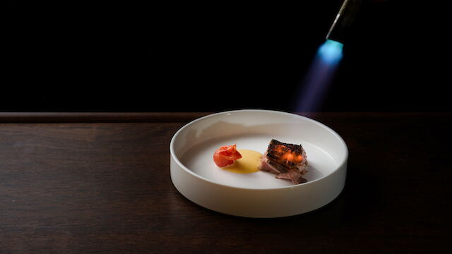 A lit torch sears a plated piece of steak with sauce and tomato on a white bowl against a dark background.