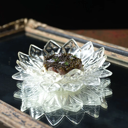 A decorative glass dish with an ornate design holds a scoop of creamy dessert topped with dark garnish, set on a reflective surface.