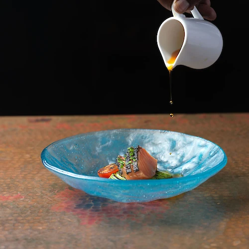 A hand pours sauce from a small white pitcher into a blue bowl containing a gourmet dish, featuring garnished vegetables on a textured surface.