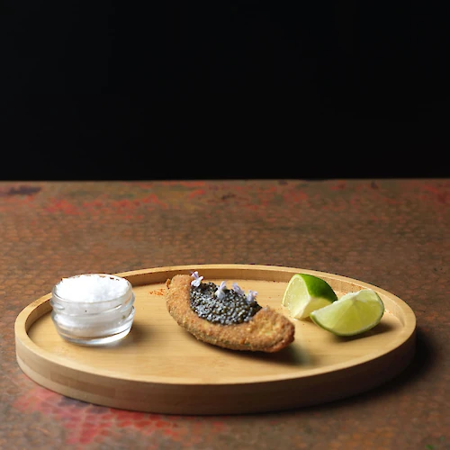 A wooden tray with a small glass jar of salt, a bread slice topped with caviar, and lime wedges, placed on a table in a dark setting.