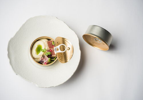 Open can of food with creamy contents, meat and vegetables, on a paper plate with the lid beside it.