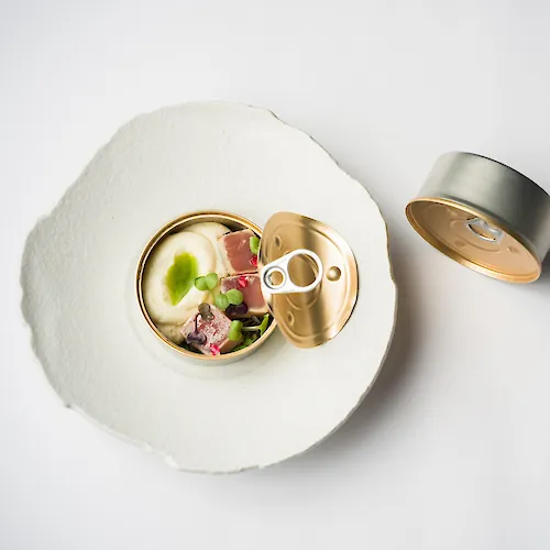 Open can of food with creamy contents, meat and vegetables, on a paper plate with the lid beside it.