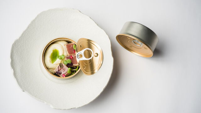 Open can of food with creamy contents, meat and vegetables, on a paper plate with the lid beside it.