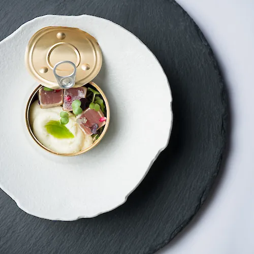 A gourmet starter served in an opened can on a slate plate, with ham, creamy sauce, microgreens, and edible flowers.