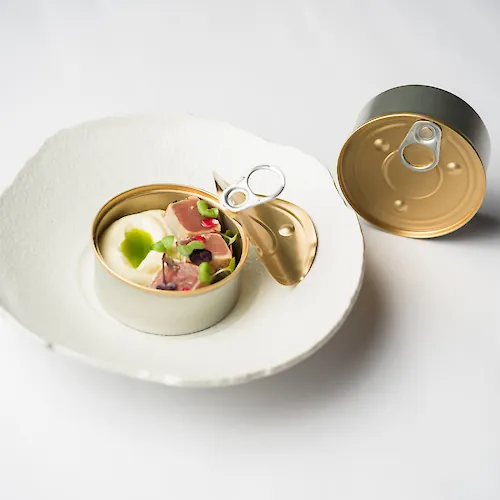 An open can of food with meat and vegetables, beside a closed can on a plate.