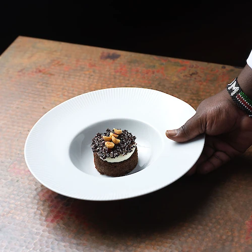 A hand is holding a white plate with a chocolate dessert garnished with nuts on a textured brown surface.