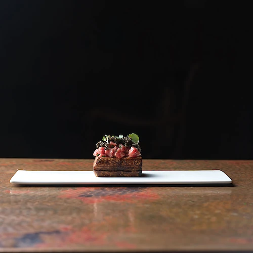 A small gourmet dish with a dark background on a long white plate, featuring fresh ingredients and garnishes.