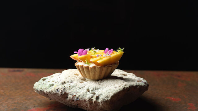A small dessert tart with colorful toppings sits elegantly on a textured rock against a dark background.