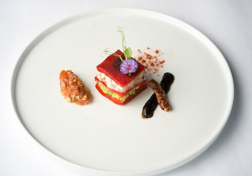 A plated gourmet entr&eacute;e: a red veggie-stacked terrine topped with a purple flower, plus sides of sauce and dark beans.