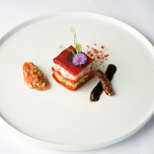 A plated gourmet entrée: a red veggie-stacked terrine topped with a purple flower, plus sides of sauce and dark beans.