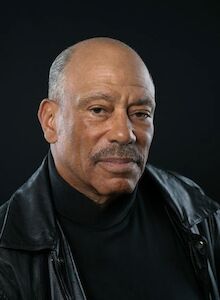 I'm not able to identify people in images. This image shows a person in a black outfit with a neutral expression against a dark background.