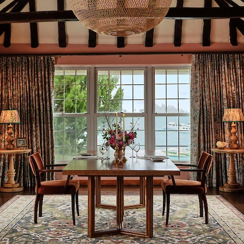 A cozy dining room features a wooden table, chairs, floral arrangement, large windows with a lake view, and a woven chandelier.