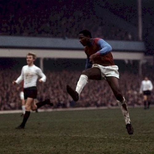 A soccer player in claret and blue kit is mid-air during a match, with another player in white and black in the background.