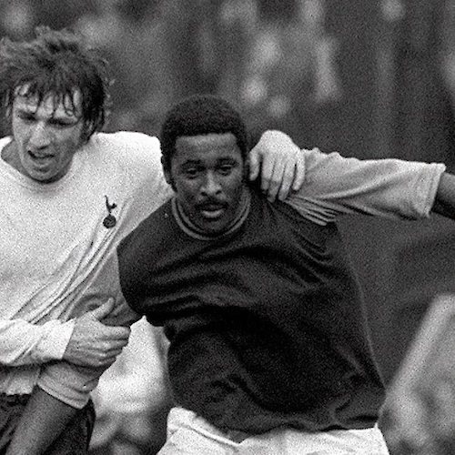 Two soccer players are in action on the field; one in a white jersey and the other in a dark jersey during a match.