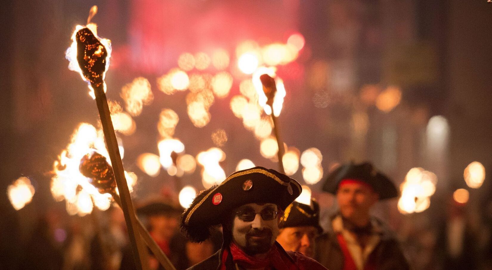 People in historical costumes holding torches march through a dimly lit street, with others blurred in the background.