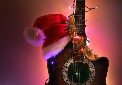 A guitar adorned with Christmas lights and tinsel, wearing a Santa hat, glowing warmly in colorful ambient light.