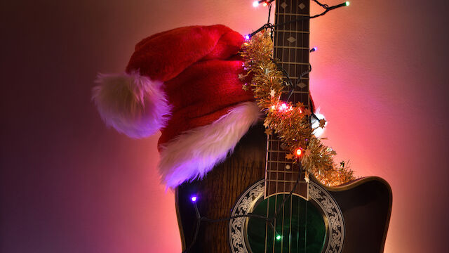 A guitar adorned with Christmas lights and tinsel, wearing a Santa hat, glowing warmly in colorful ambient light.