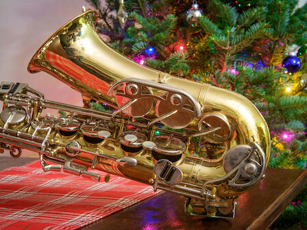 A gleaming saxophone rests on a table beside a Christmas tree adorned with colorful lights and ornaments, festive and musical.