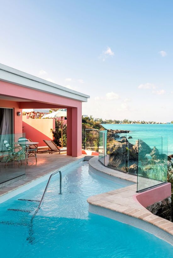 A pink seaside villa with a curved infinity pool overlooking turquoise ocean, glass railing, sunny sky, and tropical greenery.