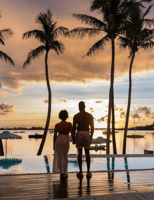 Two people stand hand-in-hand by a sunset pool, palm trees framing a tropical scene. The calm water reflects the sky as the sun dips.