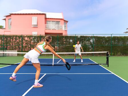 Two people play tennis on a blue court near a pink building, with a net between them and a green fence in the background.