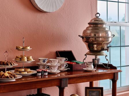 A vintage breakfast setup on a wooden sideboard against peach walls, with a large round wall clock, tea set, pastries, and a samovar by a window.