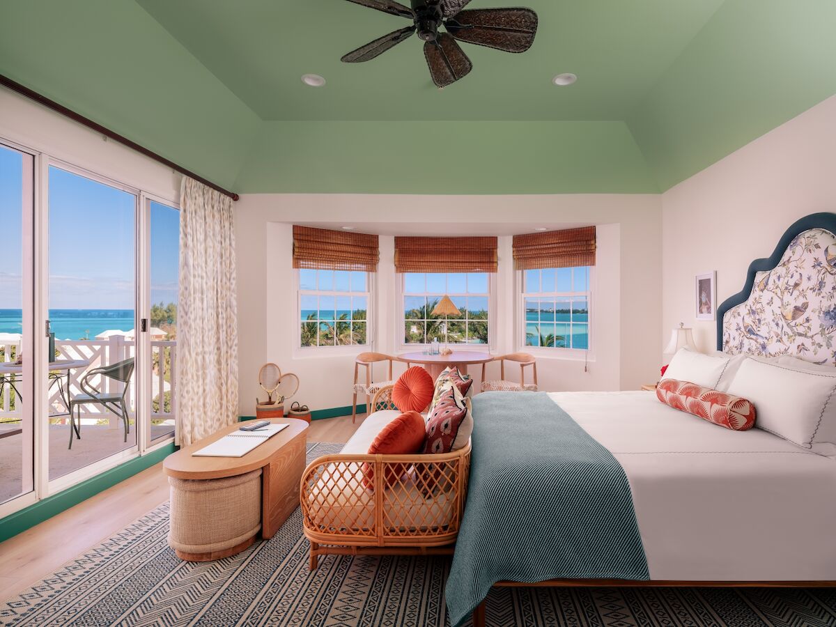 Luxurious coastal hotel room with a king bed, teal accents, wicker seating, and a balcony overlooking the ocean. A relaxing beachfront retreat.