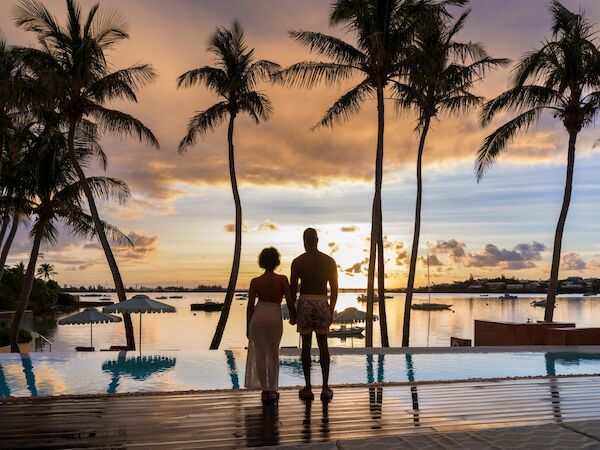 Two people stand on a wooden deck by a pool at sunset, palm trees framing the scene, watching the calm water and distant boats.