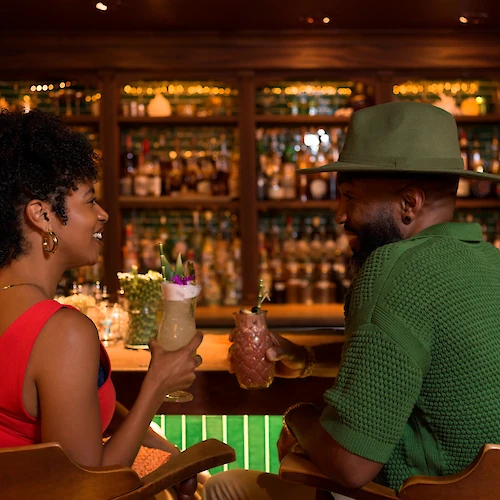 Two friends chat at a bar, sharing cocktails and smiles as they enjoy a warm, intimate evening.