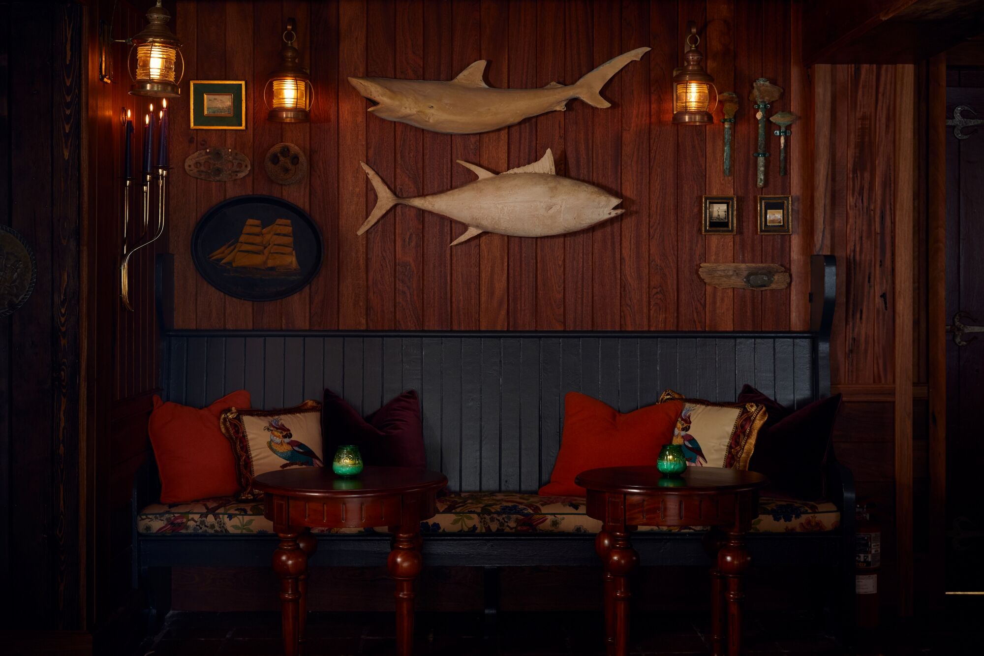 A cozy, dimly lit sitting area with wooden walls, two red-cushioned chairs, a blue bench, and framed marine decor including three mounted fish on the wall.